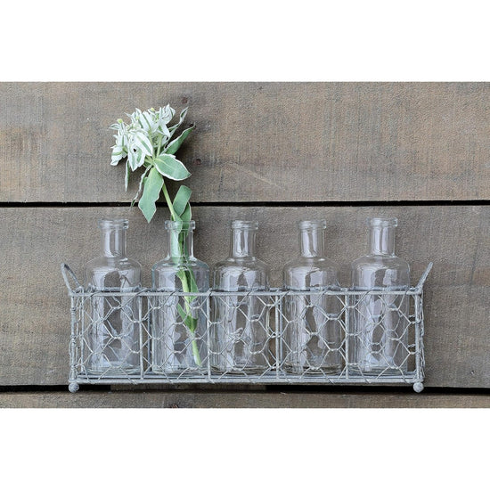 Set of glass bottles in a metal basket with a single white flower against a wooden background