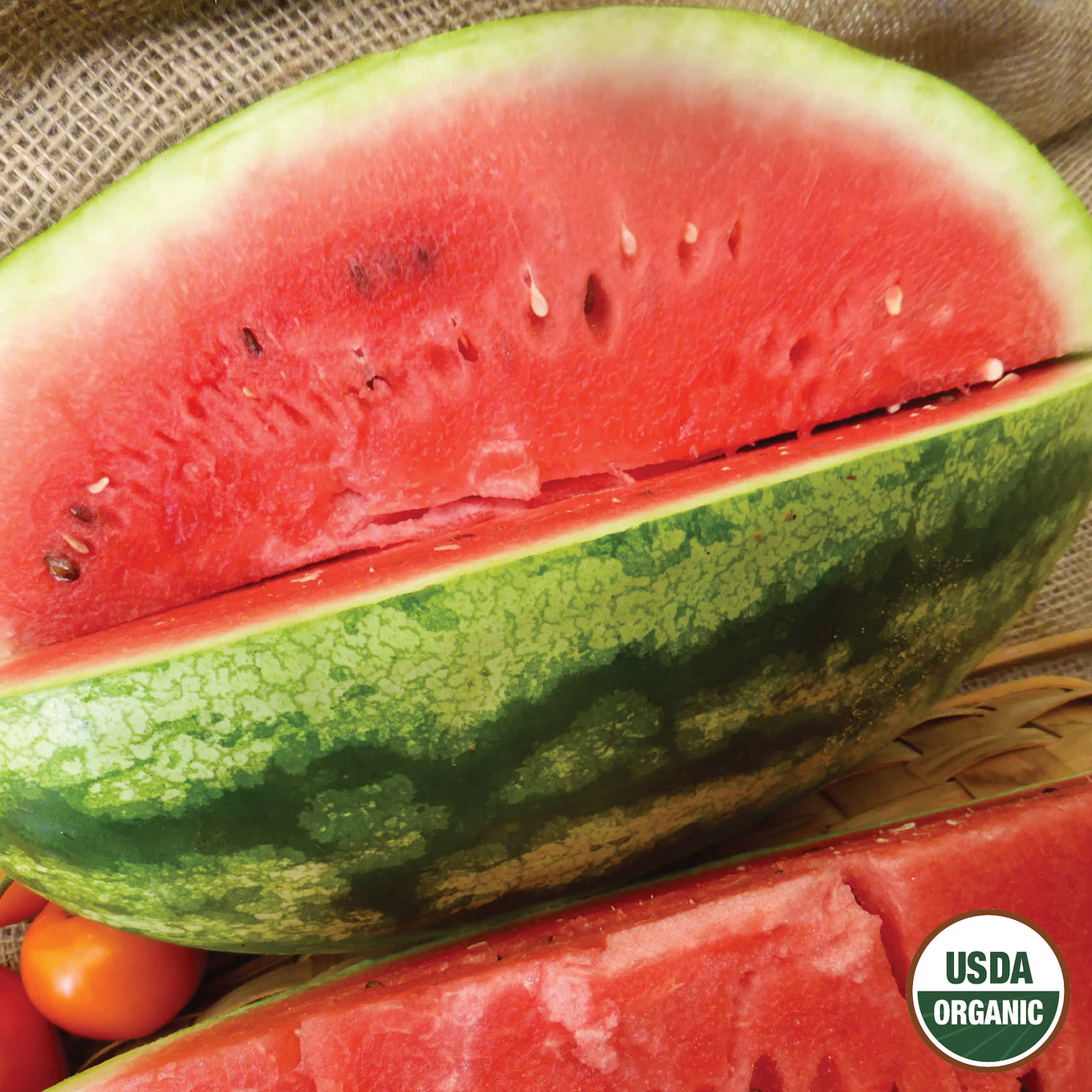 A close-up of a Crimson Sweet organic watermelon cut in half, displaying the deep red flesh and the green striped rind, with a USDA Organic label in the corner.