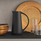Black pitcher on a dark surface with a woven plate and glasses in the background.