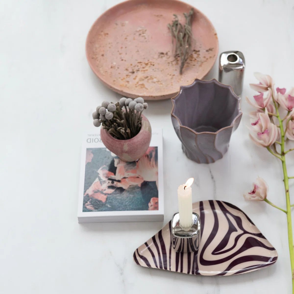 Ceramic items including a plate, planter, and candle holder on a white surface with flowers and a book.
