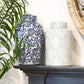 Two decorative vases on a mantelpiece with a plant and wall art in the background.