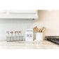 Kitchen counter with a bread bin, utensils, and flowers on a white tiled wall background.
