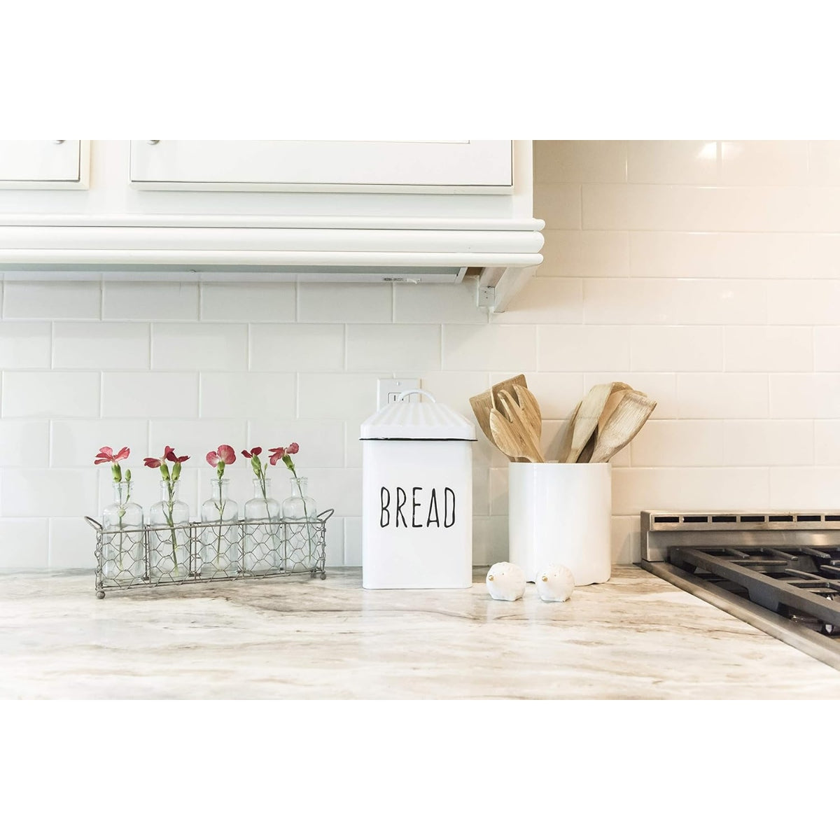 Kitchen counter with a bread bin, utensils, and flowers on a white tiled wall background.