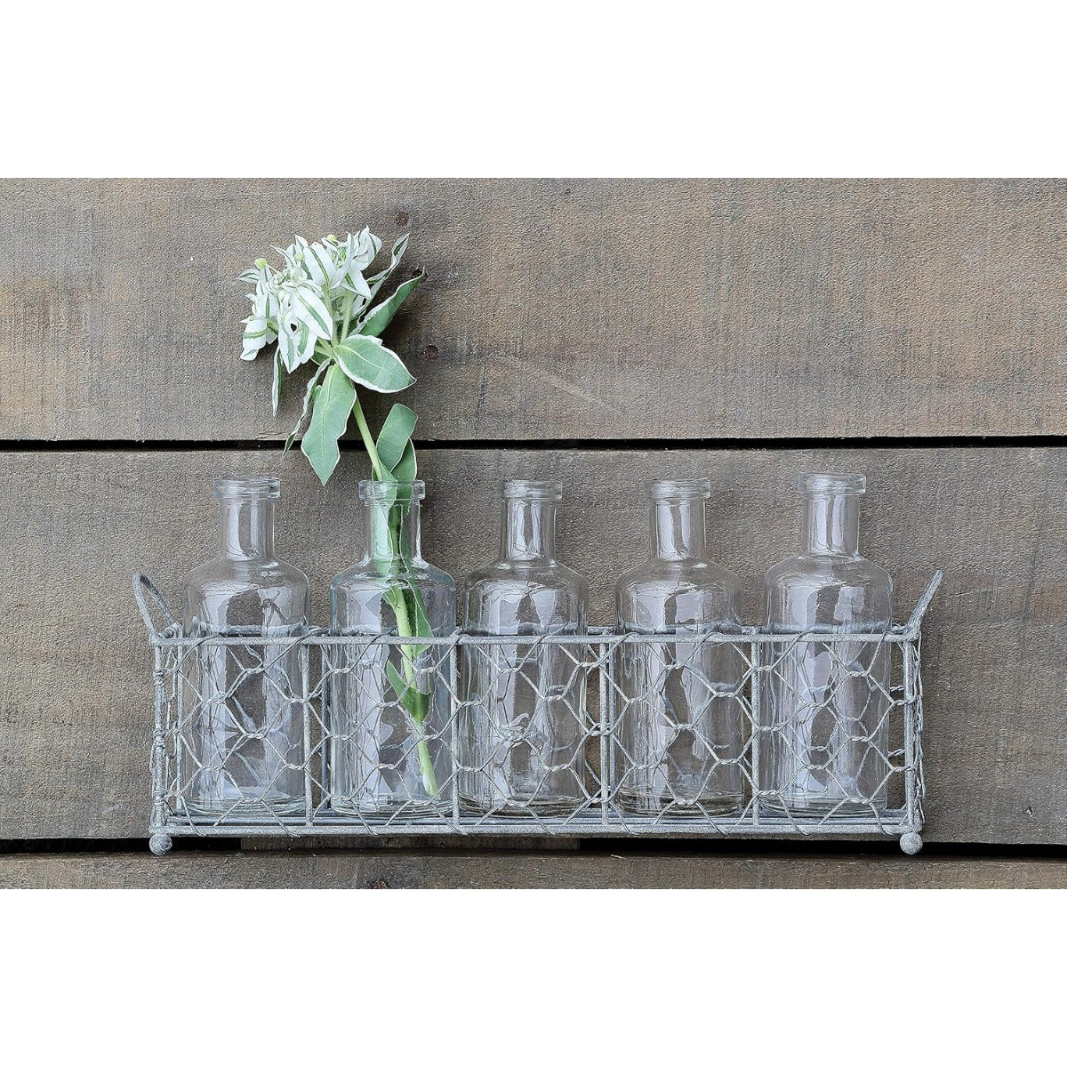Set of glass bottles in a metal basket with a single white flower against a wooden background