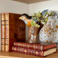 Decorative bird figurine on a stack of books with a vase in the background