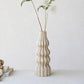 A tall, textured vase in a warm off-white tone, styled with white flowers on a light gray surface against a neutral background.