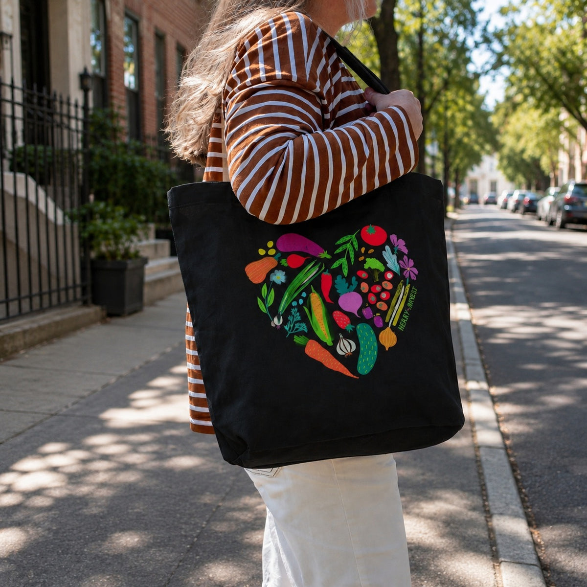 A person carrying a black canvas tote bag on a sunny city street. The bag features a colorful heart-shaped graphic made of various illustrated vegetables and fruits, including carrots, cucumbers, peppers, and tomatoes.