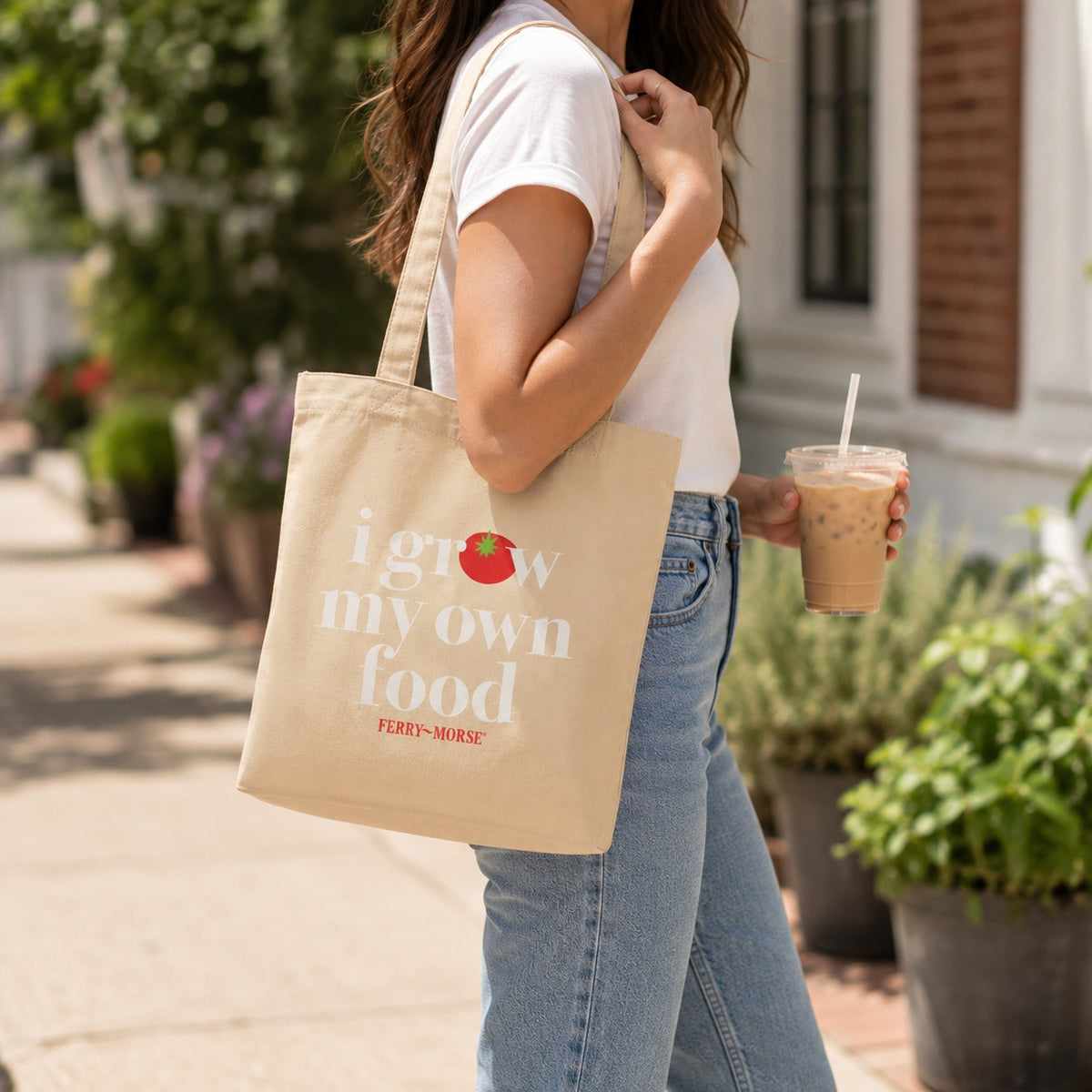 A woman wearing a white t-shirt and blue jeans carries a beige canvas tote bag with the text 'i grow my own food' and a small tomato graphic. She is holding an iced coffee on a sunny outdoor sidewalk.