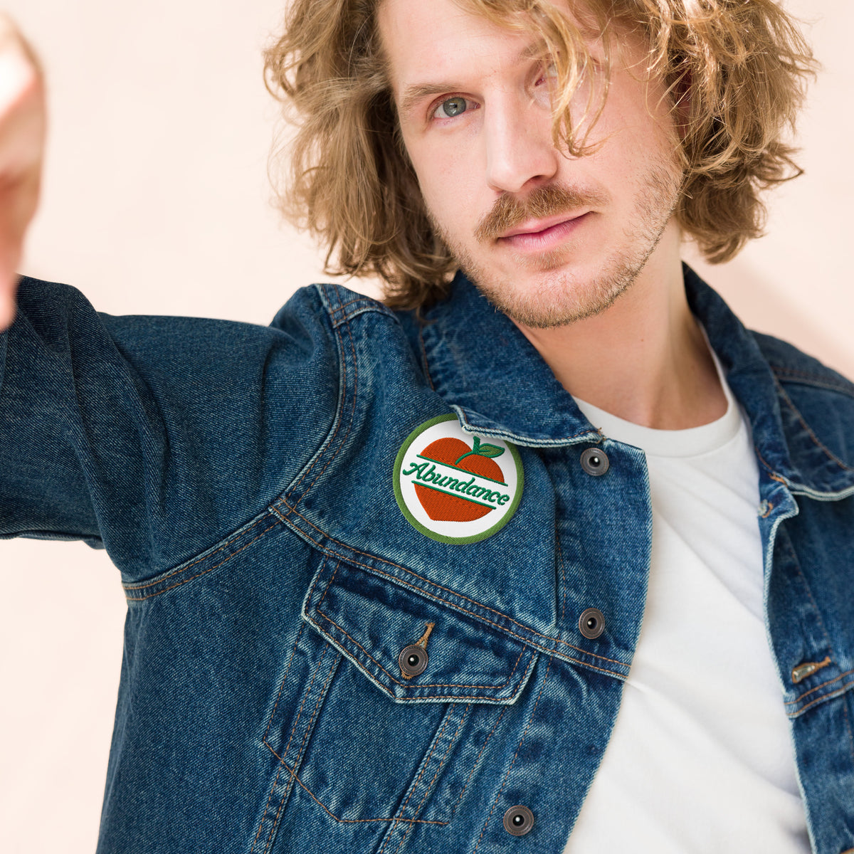 Man wearing a denim jacket with an 'Abundance' patch on a beige background