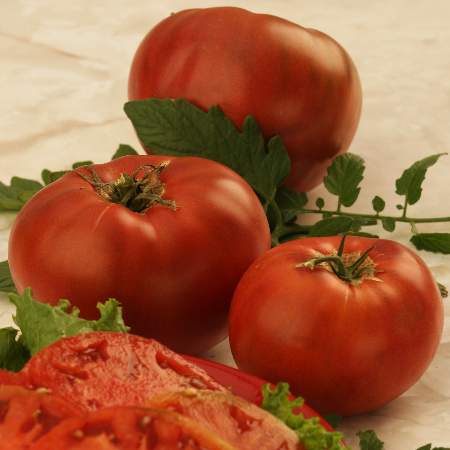 Cherokee Purple Tomatoes sitting on a table next to more Cherokee Tomato that has been sliced. These Cherokee Purple Tomatoes are a purple heirloom tomatoes variety.