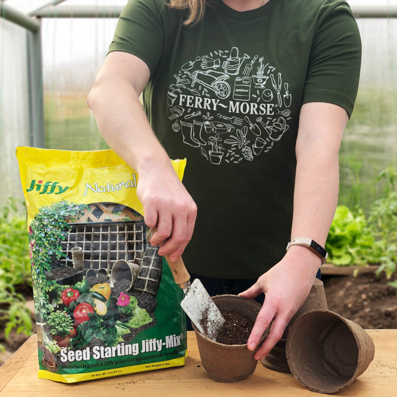 Female gardener wearing Ferry-Morse logo t-shirt in Size L and adding Seed Starting Jiffy Mix to Jiffy Peat Pots in a greenhouse